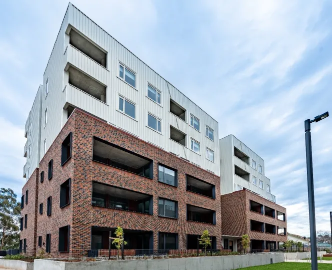 A wide view of a white and brick apartment building with landscaped garden beds, open green space and paved walkways in front.