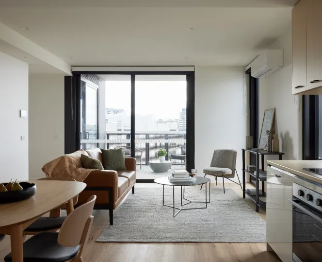A brightly lit apartment showing the kitchen, dining and lounge room. The large glass doors at the back lead onto a balcony overlooking city views.