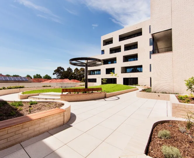 An outdoor paved courtyard with gardens beds, seating areas and open green space. The corner of a large apartment building sits behind it. 