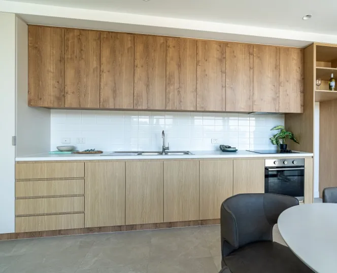 A close-up image of the kitchen cabinetry, featuring a sink, stovetop, oven, and ample bench space, with a dining table visible to the right. 