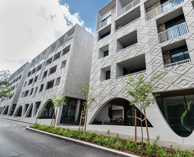 An image of textured concrete façade of the buildings featuring archways and balconies, facing Kirner Street, with greenery lining the kerb. 