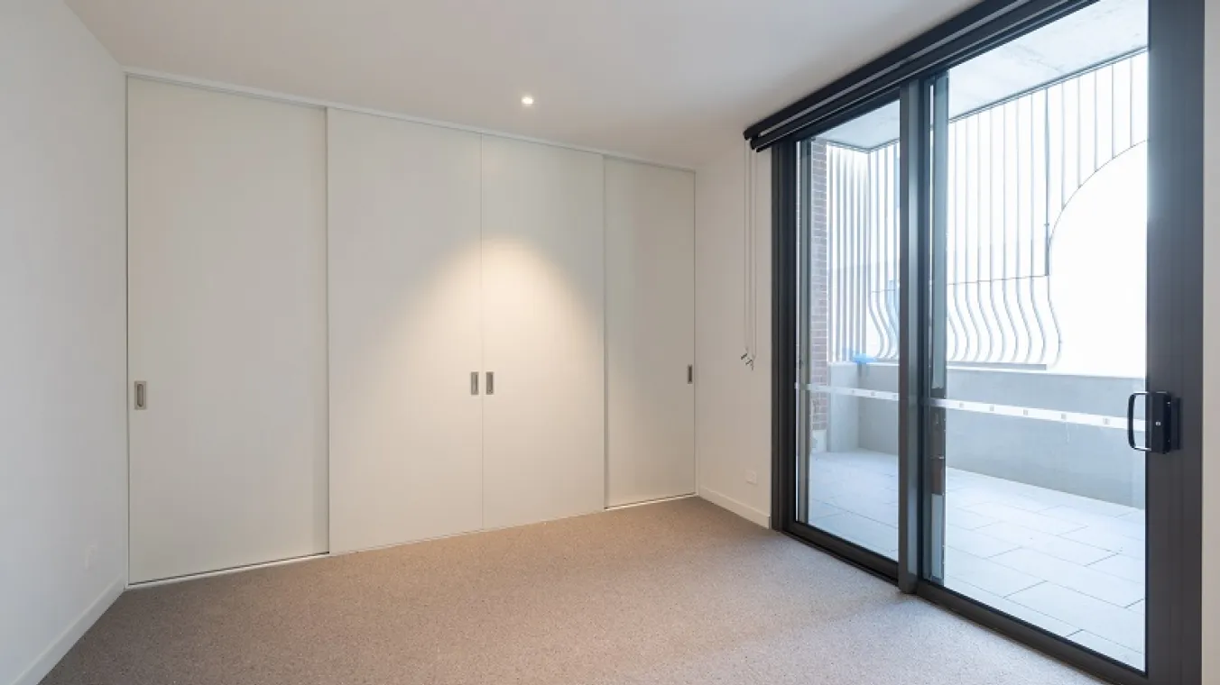Image of bedroom with carpet, built-in wardrobe, and a transparent glass sliding door in the room with natural light.