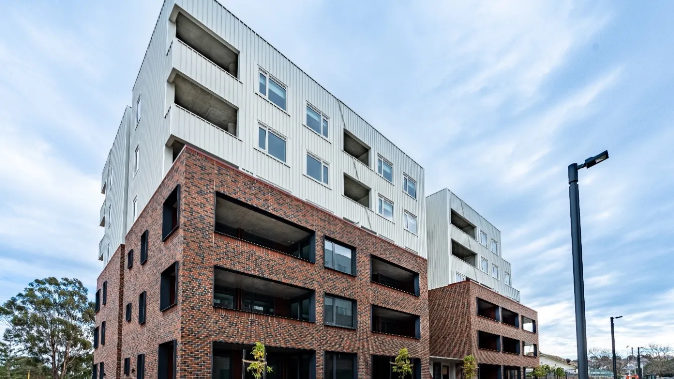 A wide view of a white and brick apartment building with landscaped garden beds, open green space and paved walkways in front.
