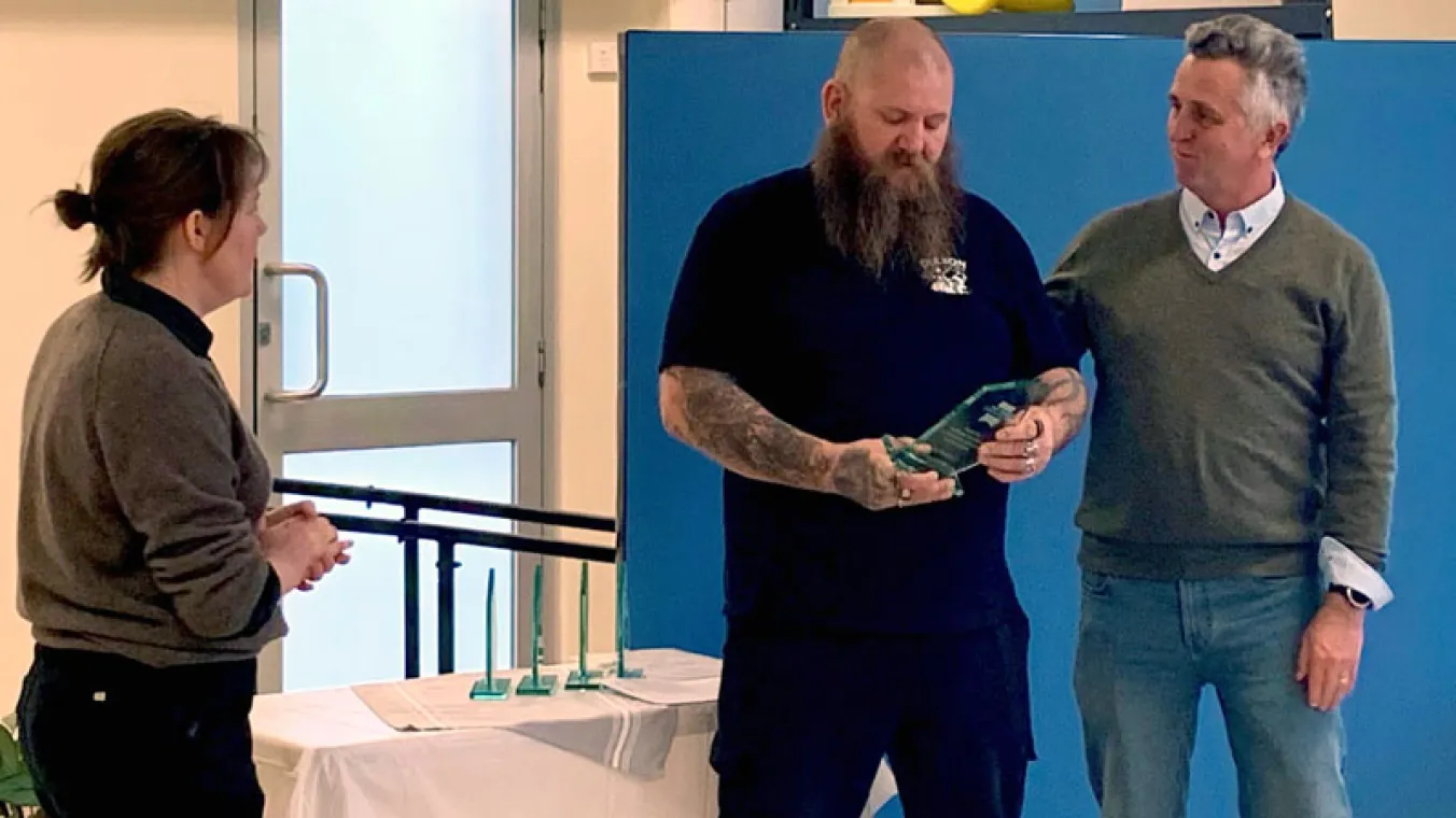 South Division Housing Manager, Paul Bolt, congratulates Park Towers Food Pantry founder and award winner, Troy Matthews, along with Park Towers Community Engagement Manager, Lizzy Harding. The three are standing in front of a table with awards. Troy is holding an award.