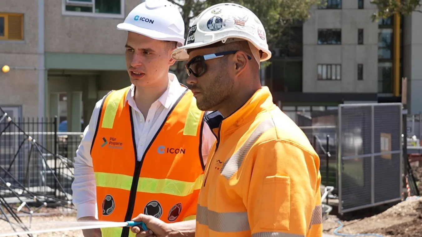 Image of 2 men wearing orange hi-vis vests spraying water at a construction site.    