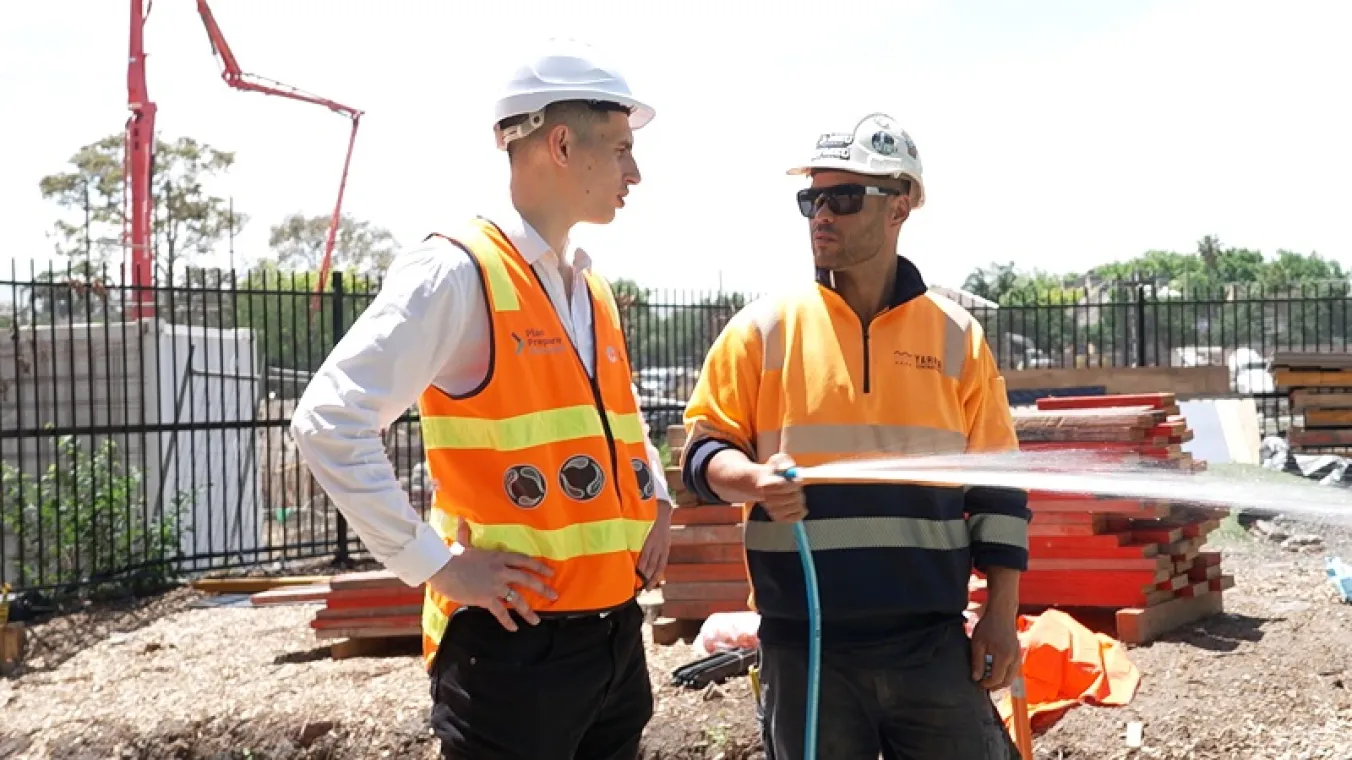 Image of 2 men wearing orange hi-vis vests spraying water and discussing at a construction site.      