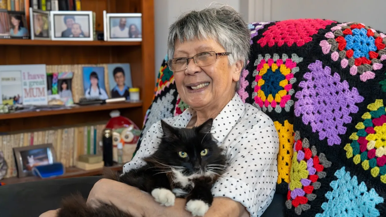 Image of a old lady sitting on couch while holding her black cat in her arms.  