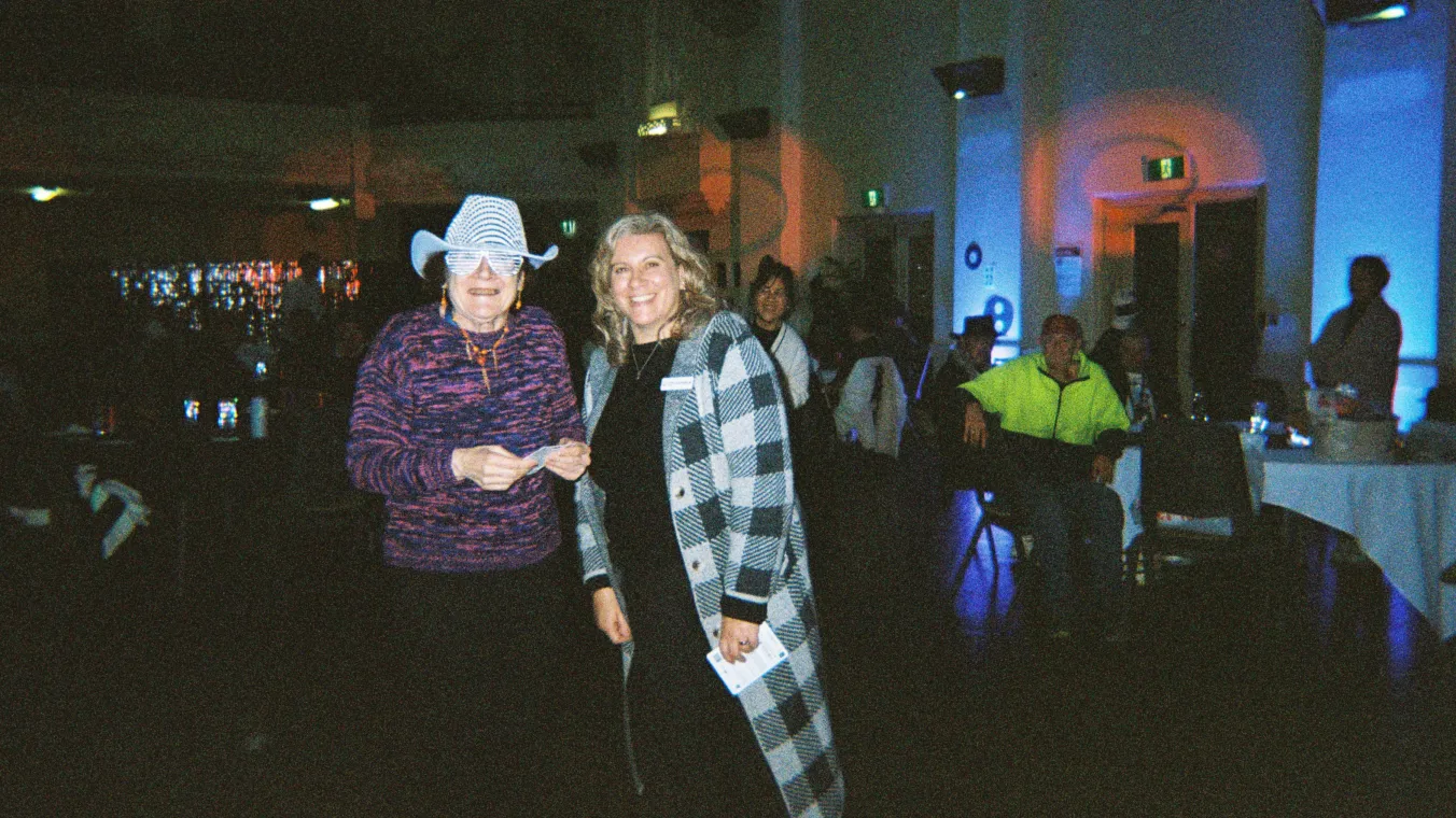 2 women looking at the camera smiling. The woman on the left is wearing a cowboy hat and novelty glasses