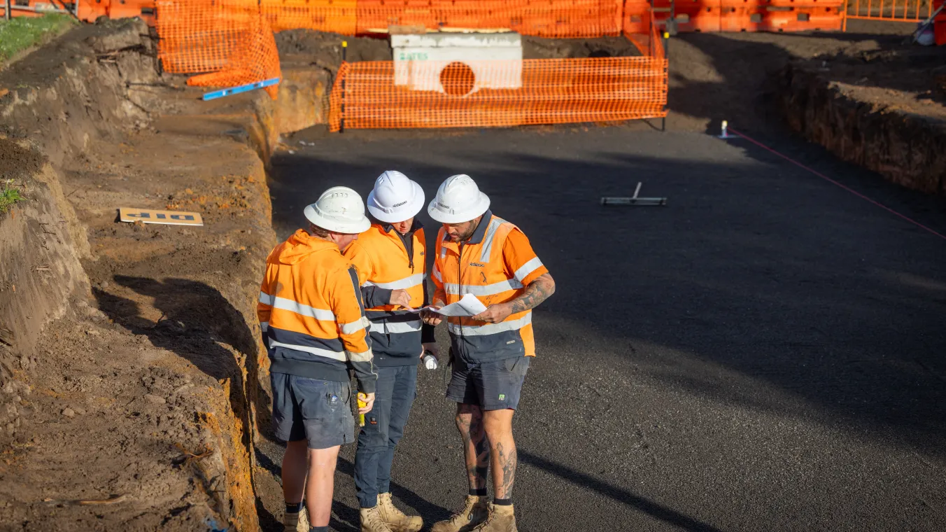 Construction workers wearing orange safety vests and white hard hats work at a construction site.